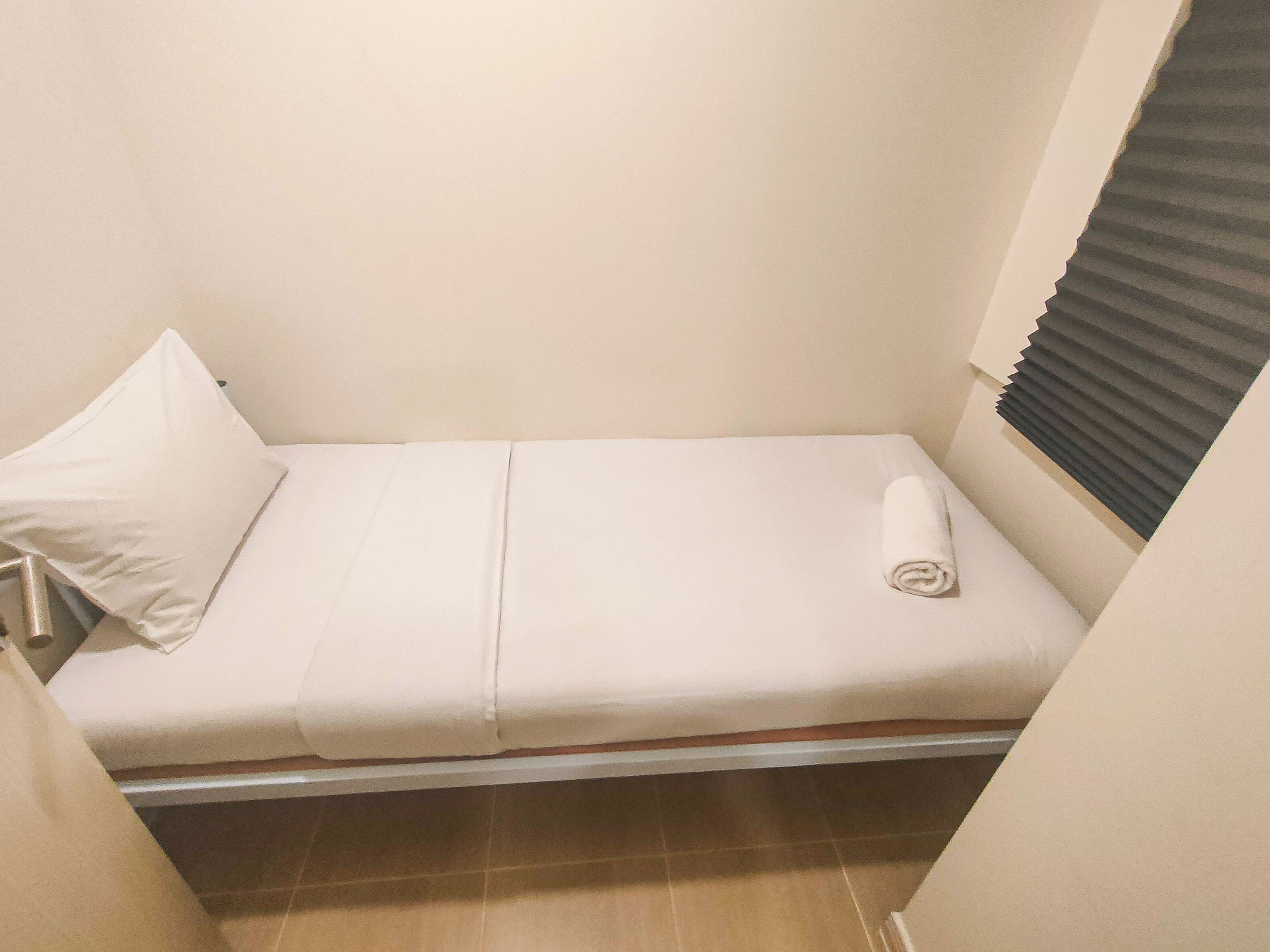 A small, single-bed room with a white bedspread, a rolled towel, and a pillow.