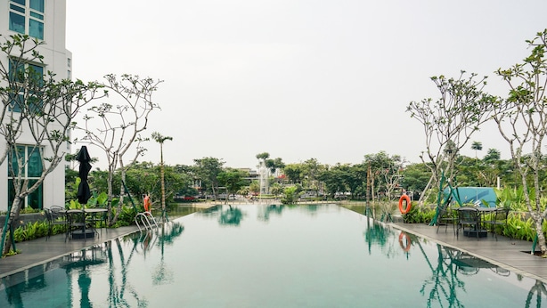 A swimming pool with surrounding greenery and a modern building in the background. - Cozy And Enjoy Living 1Br At Citralake Suites Apartment (Jakarta)