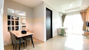 A modern living room with a dining area, a large window with curtains, a flat-screen TV, and a built-in air conditioning unit. - Cozy And Enjoy Living 1Br At Citralake Suites Apartment (Jakarta)