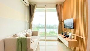 A modern living room with a sofa, a TV mounted on a wooden panel, and a large window with curtains. - Cozy And Enjoy Living 1Br At Citralake Suites Apartment (Jakarta)