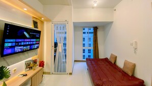 A modern living room with a flat-screen TV mounted on the wall, a red sofa, a wooden bench, and a small table with a vase and a book. - Nice View And Cozy 1Br Apartment At Tokyo Riverside Pik 2 (Teluknaga)