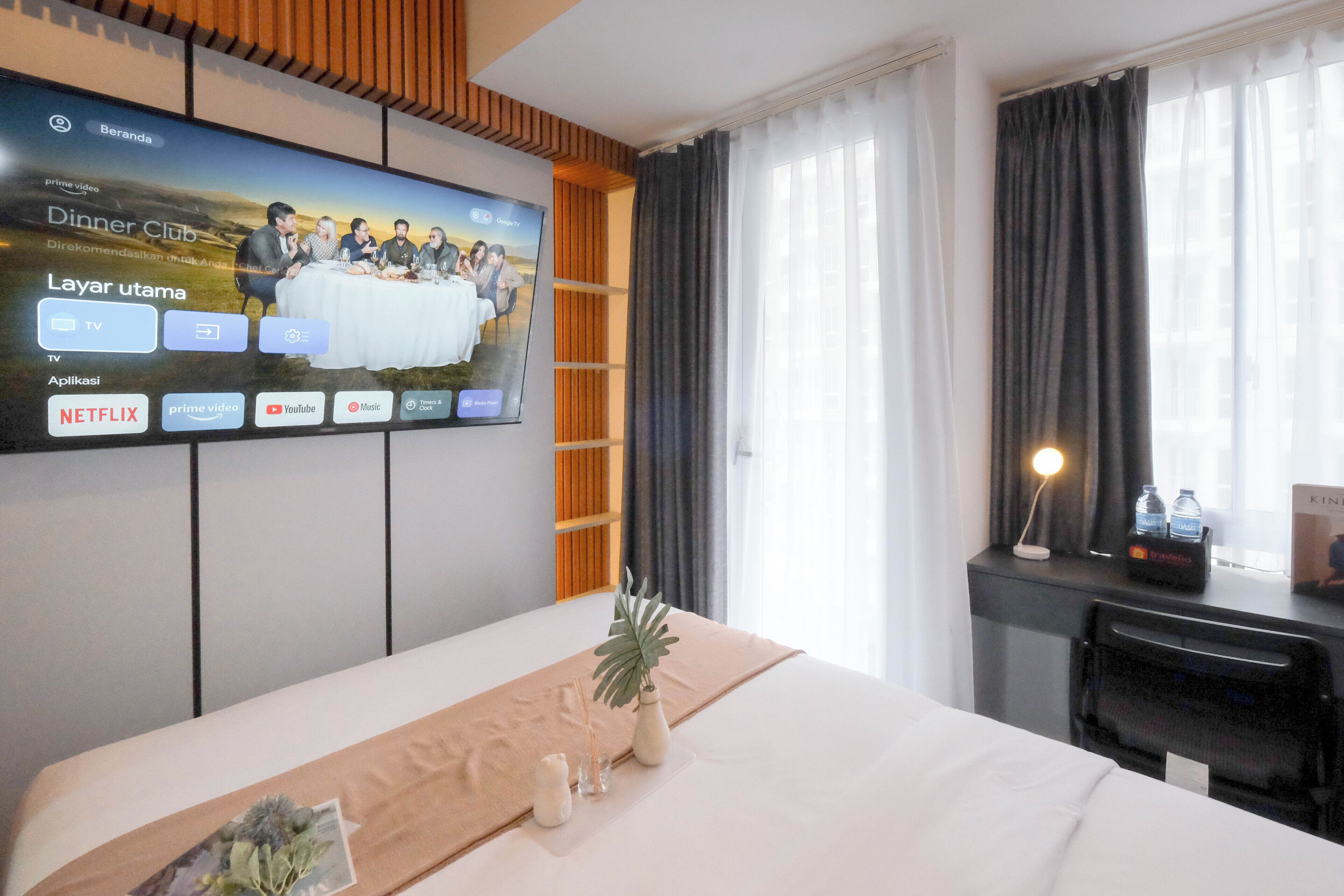 Ein Hotelzimmer mit Bett, einem an der Wand montierten Fernseher, einem Schreibtisch mit Lampe und einem Fenster mit Vorhängen.