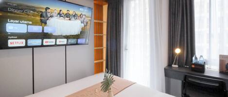 Ein Hotelzimmer mit Bett, einem an der Wand montierten Fernseher, einem Schreibtisch mit Lampe und einem Fenster mit Vorhängen.
