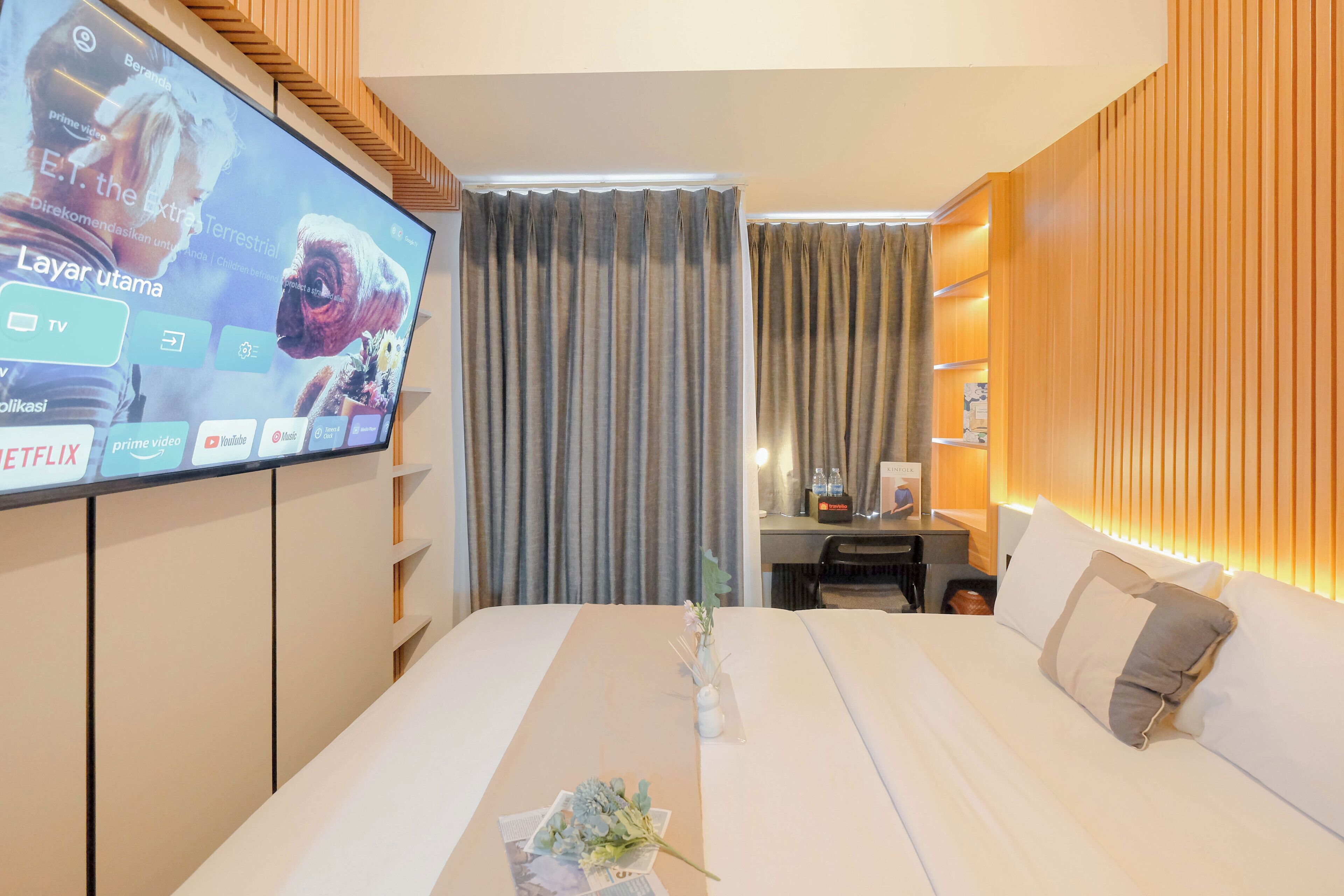 Habitación de hotel con un televisor grande en la pared, cama con ropa blanca, escritorio con lámpara y paneles de madera en las paredes.