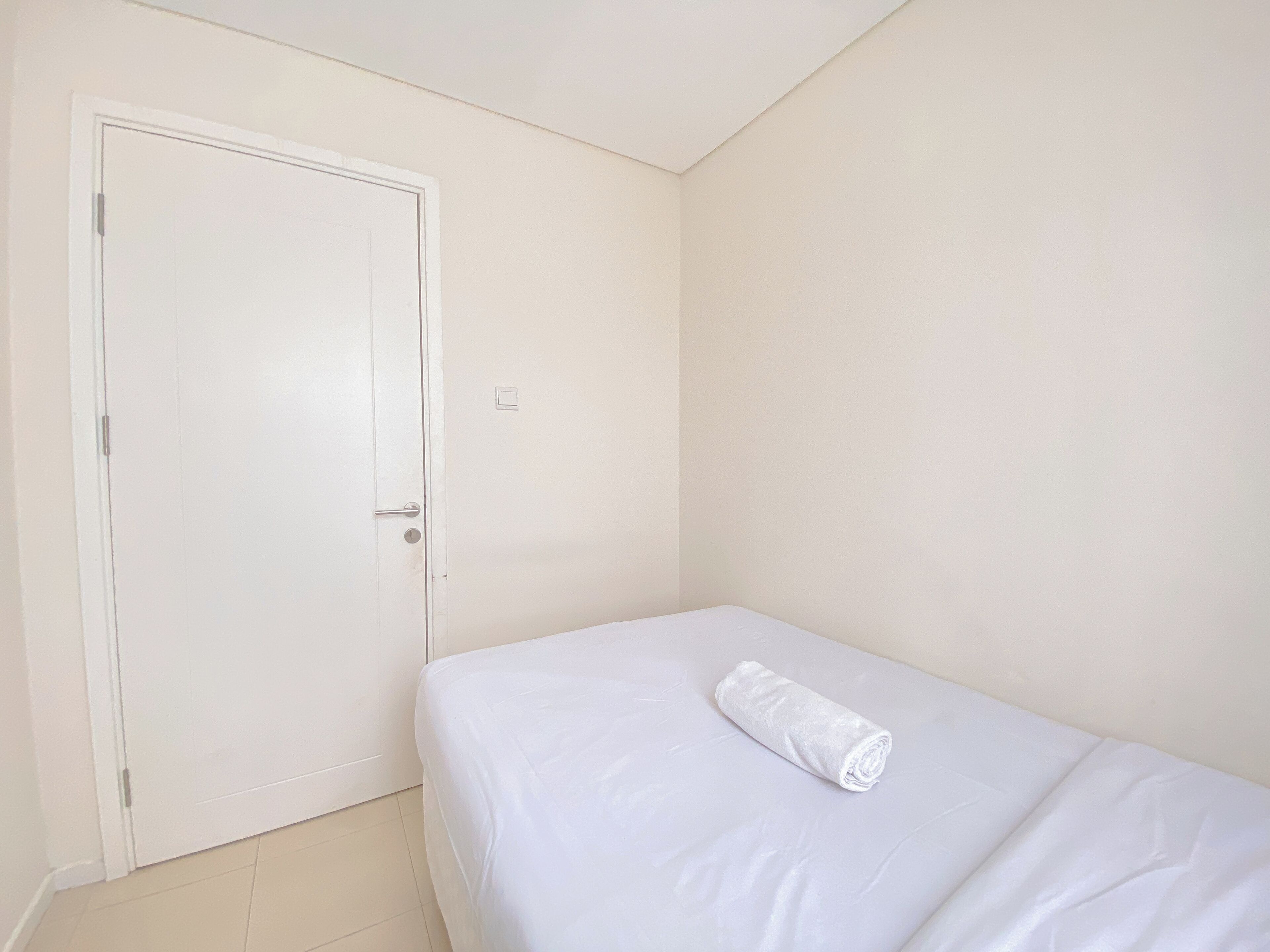 A small, clean room with a single bed, a white door, and a light switch on the wall.