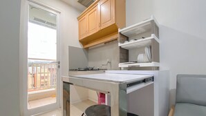 A compact kitchen with a white countertop, two black stools, wooden cabinets, and a view through a glass door to an outdoor balcony. - Simply And Cozy 2Br Apartment At Parahyangan Residence (Bandung)