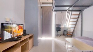 A modern, well-lit room with a flat-screen TV on a wooden shelf, a small desk with a chair, a cushioned bench, and a metal ladder against the wall. - Fully Furnished Studio With Mezzanine Bed At Serpong Garden Apartment (Cisauk)