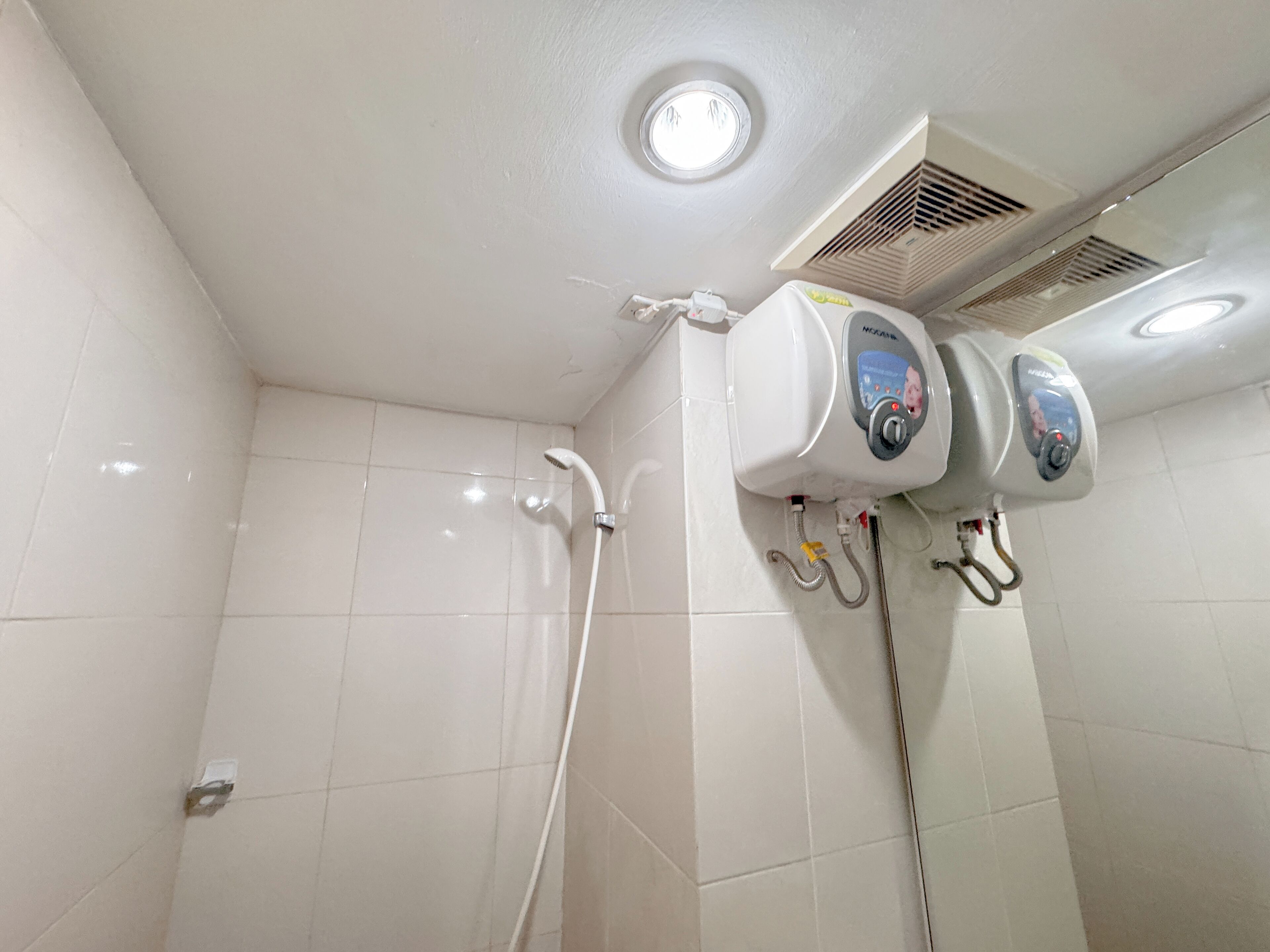 Une salle de bain avec des murs recouverts de carreaux blancs, un espace douche et un plafond doté d’un luminaire et d’une grille d’aération.