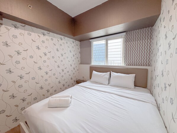 Une chambre à coucher avec un lit, du linge de lit blanc, un papier peint à motifs floraux, une fenêtre avec des stores et une vue sur des immeubles.
