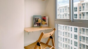 Una esquina de una habitación con una silla de madera, un pequeño estante con un libro y flores, y una ventana que ofrece vistas a edificios.