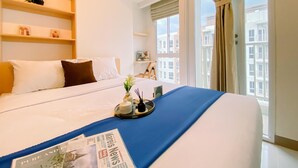 Habitación de hotel con una cama grande, una mesita de noche con una bandeja, un periódico y vistas a edificios por la ventana.