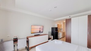 A hotel room with a bed, a desk with a chair, a television, a wardrobe, and a kitchenette. - Cozy Studio Apartment Mataram City (Ngaglik)