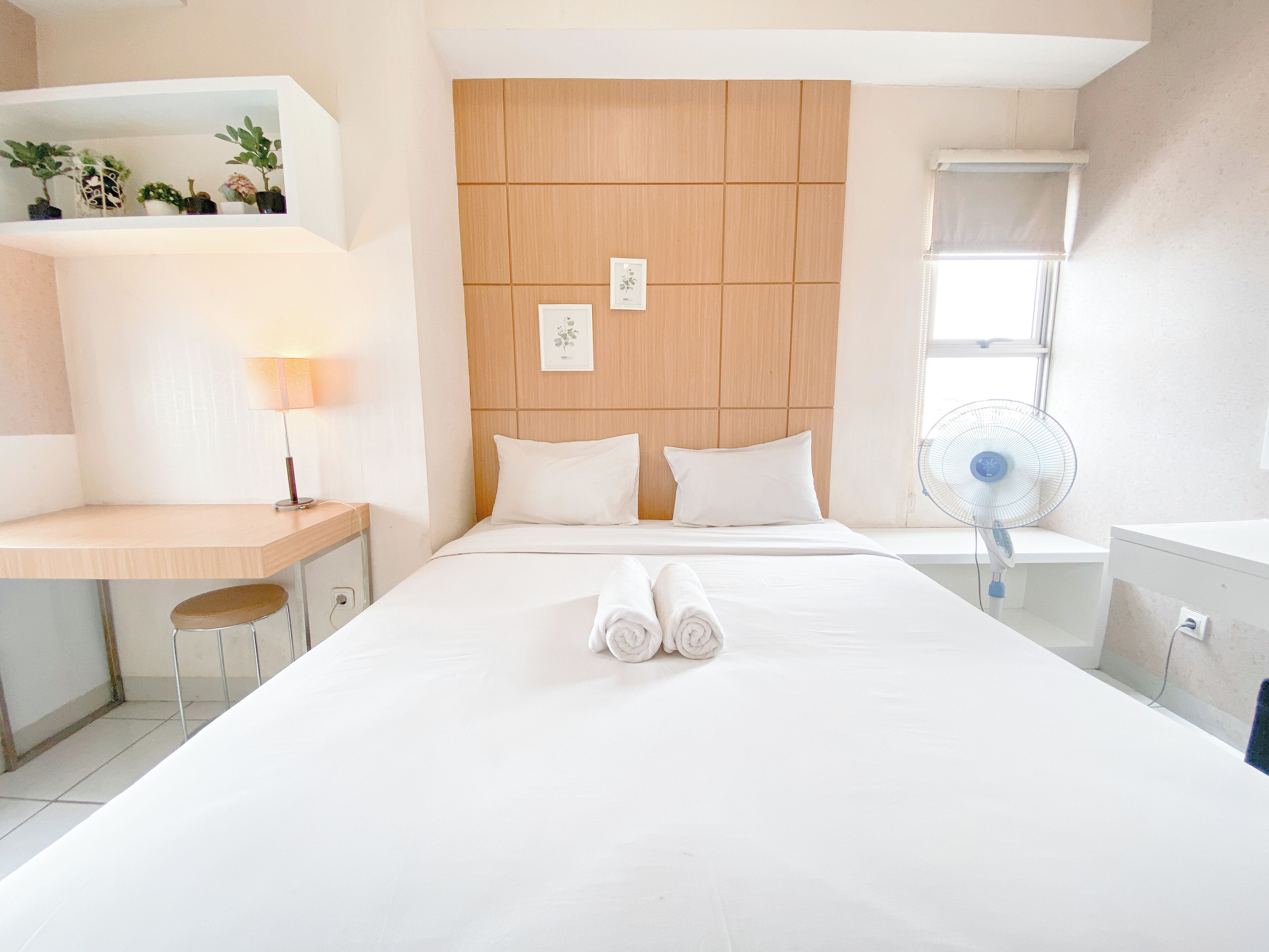A hotel room with a bed, a desk, a chair, a bedside table, a lamp, a fan, and a window.
