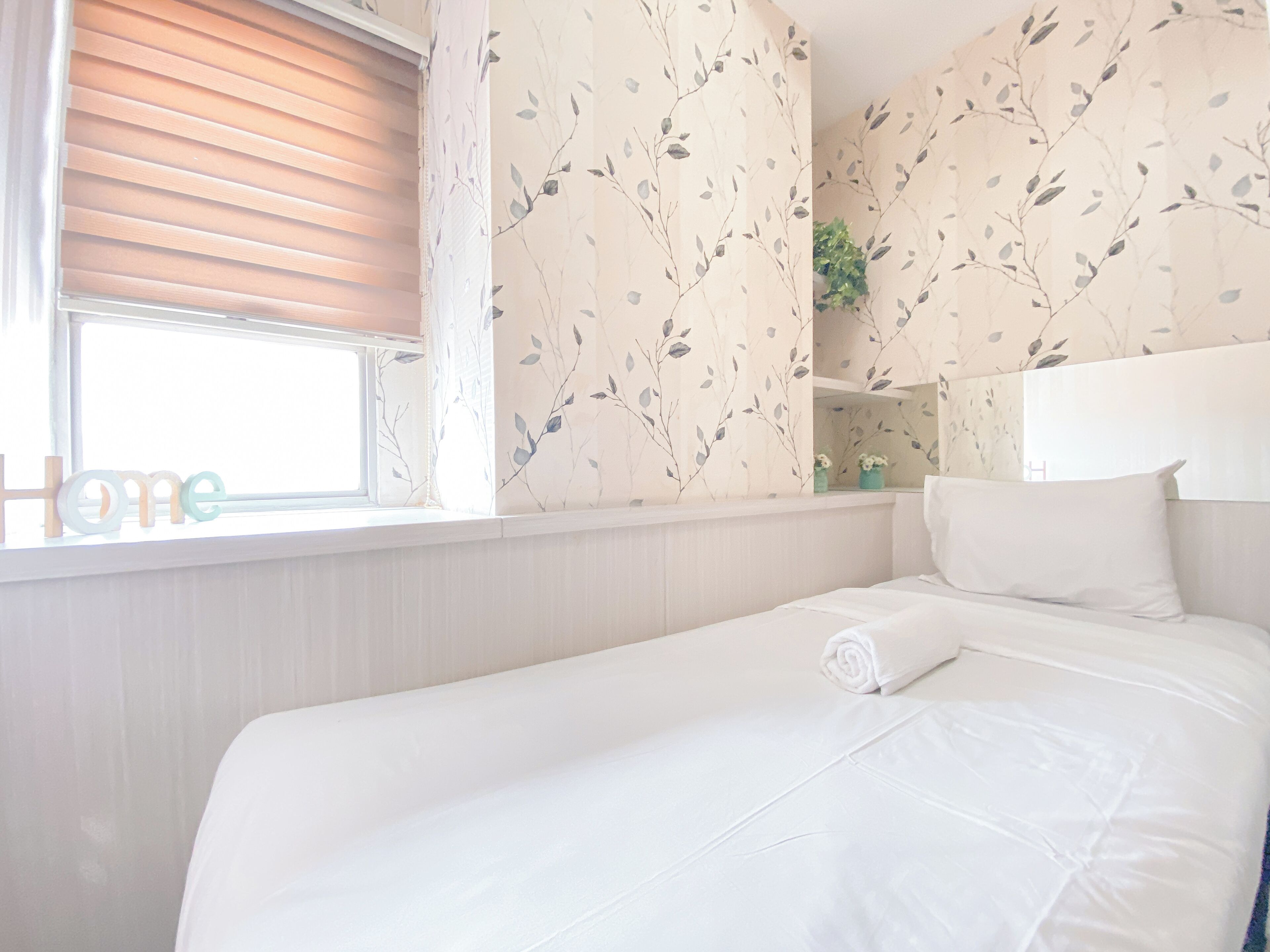 Een net opgemaakt bed met een wit hoofdeinde, een raam met zonwering en bloemenbehang.