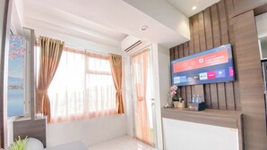 A modern hotel room with a large window, a flat-screen TV mounted on the wall, a reception desk, and a potted plant.