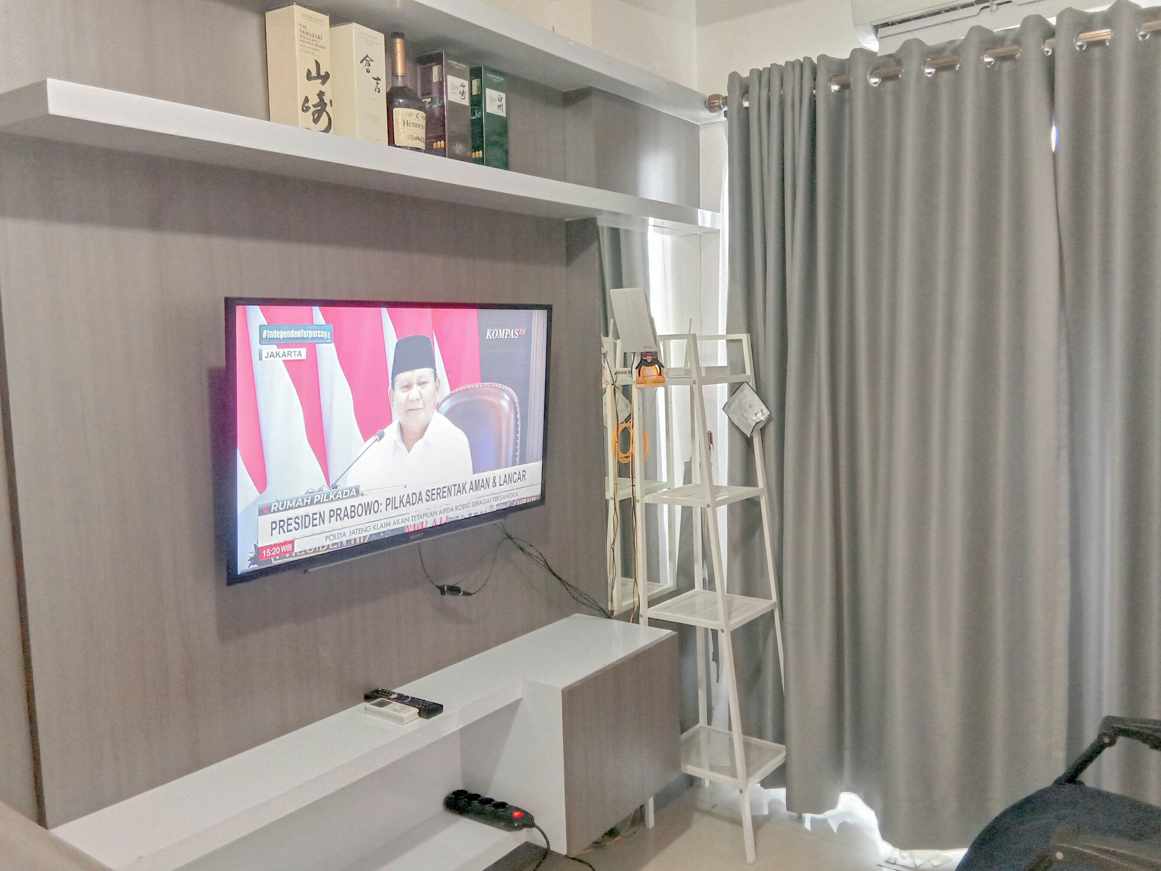 A living room with a flat-screen TV mounted on the wall, a white entertainment unit below, and a grey curtain to the right.