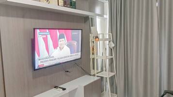 A living room with a flat-screen TV mounted on the wall, a white entertainment unit below, and a grey curtain to the right.