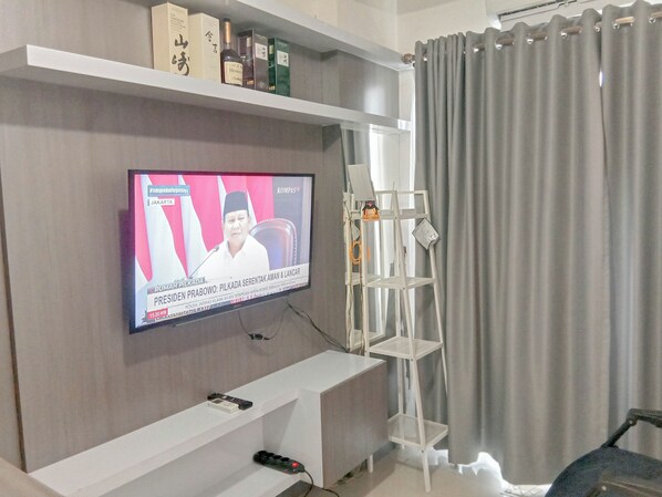 A living room with a flat-screen TV mounted on the wall, a white entertainment unit below, and a grey curtain to the right.