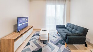 A modern living room with a flat-screen TV on a wooden stand, a grey sofa, a small white table, and a patterned floor.