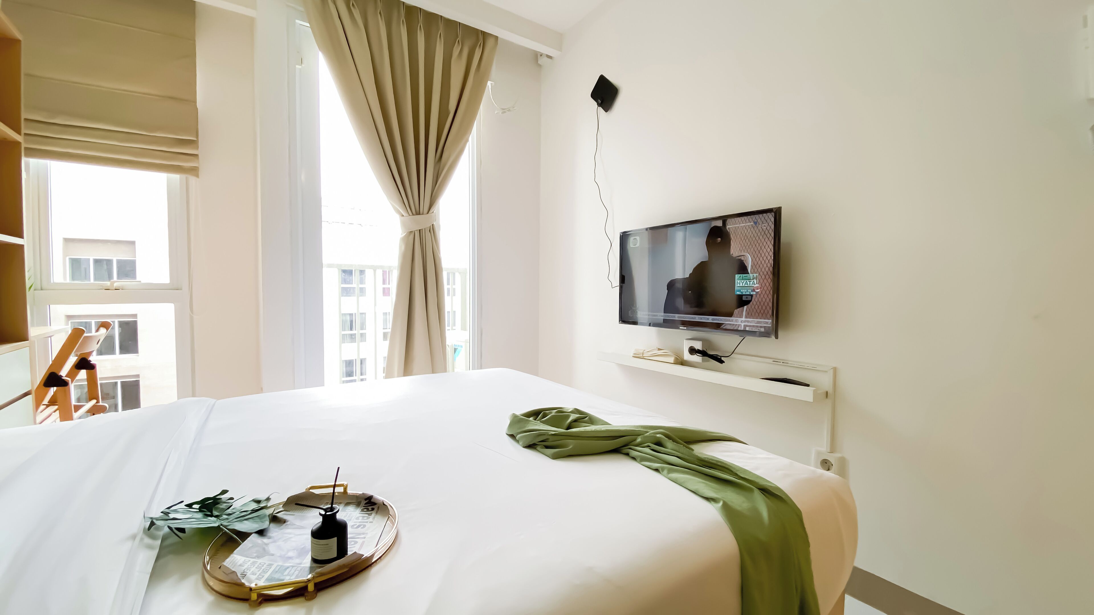 Habitación de hotel con cama, televisor montado en la pared, ventana con cortinas y vista a edificios.