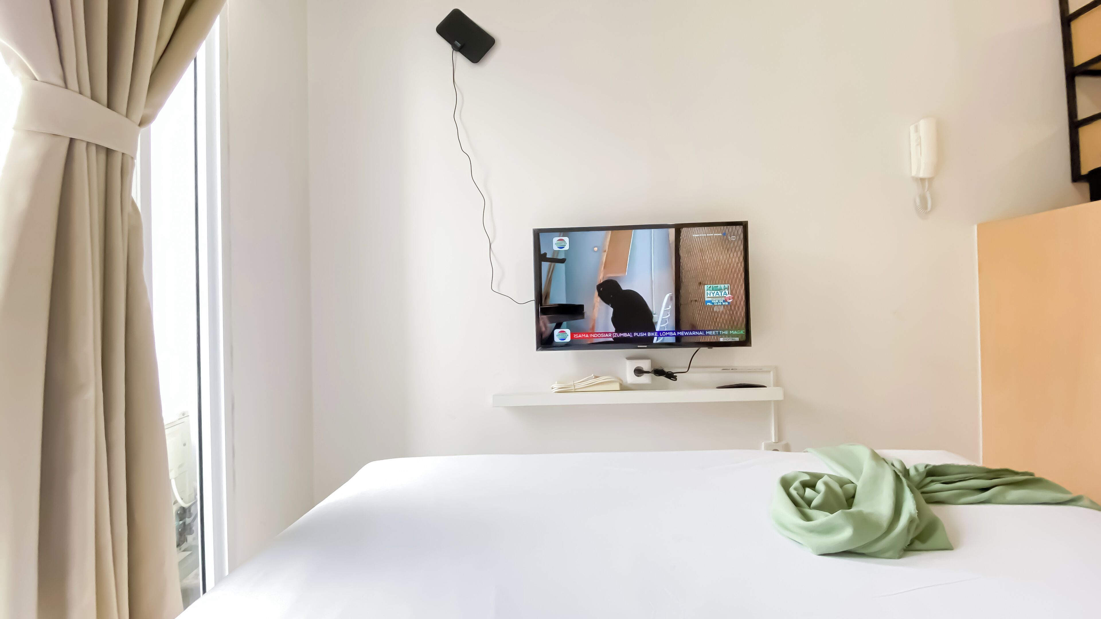 Ein Hotelzimmer mit einem Bett, einem an der Wand montierten Fernseher, einem Telefon und einem Fenster mit Vorhängen.