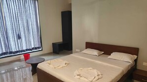 Una camera d'albergo con un letto grande, un tavolino rotondo, un secchio rosso e una finestra con tende.