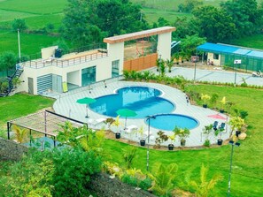 Un bâtiment moderne doté d’un espace piscine comprenant une piscine circulaire et une piscine rectangulaire séparée, le tout entouré d’un espace de détente extérieur et de verdure.