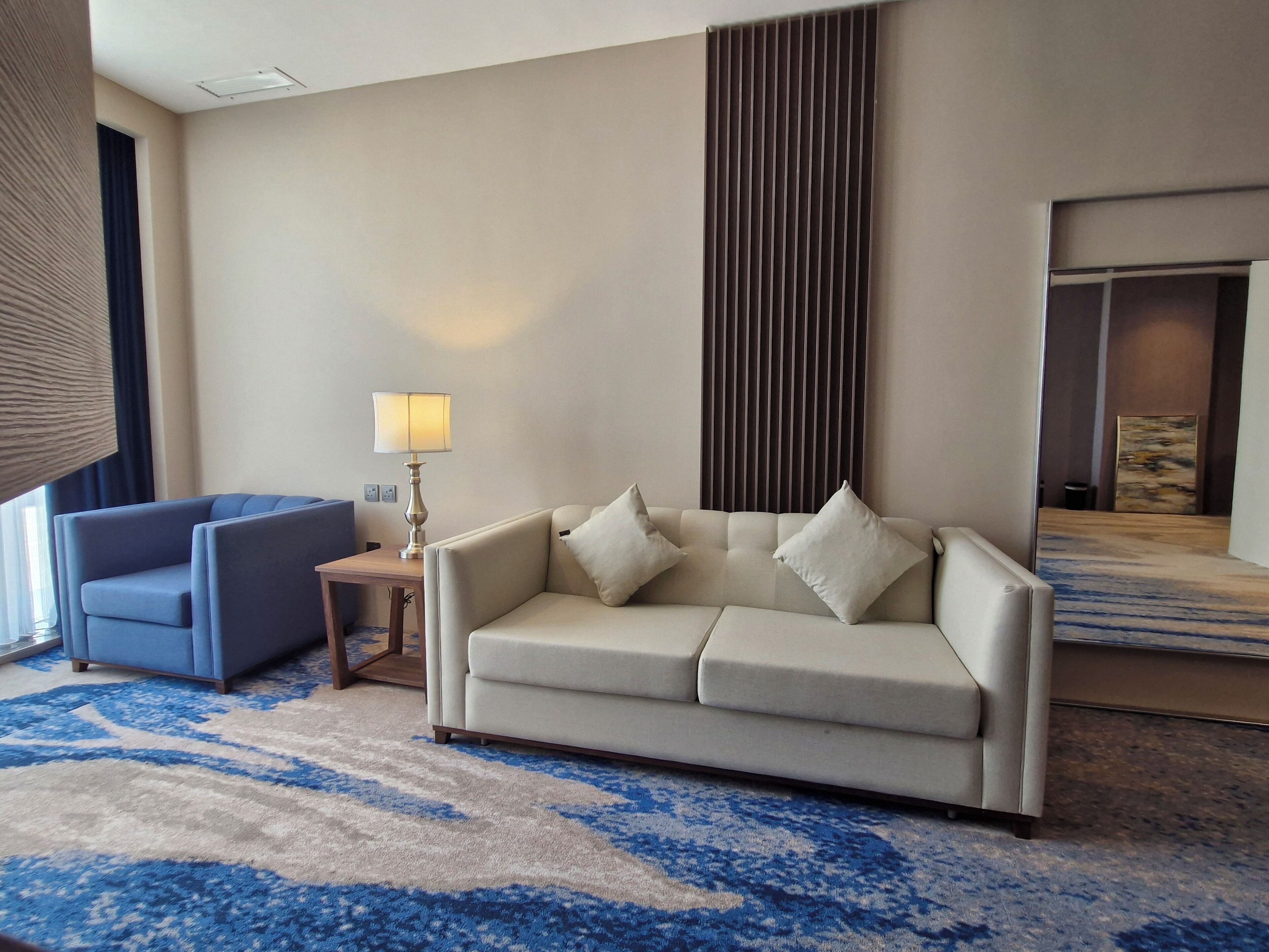 A hotel room with a beige sofa, a blue armchair, a small table with a lamp, a large mirror, and a patterned carpet.