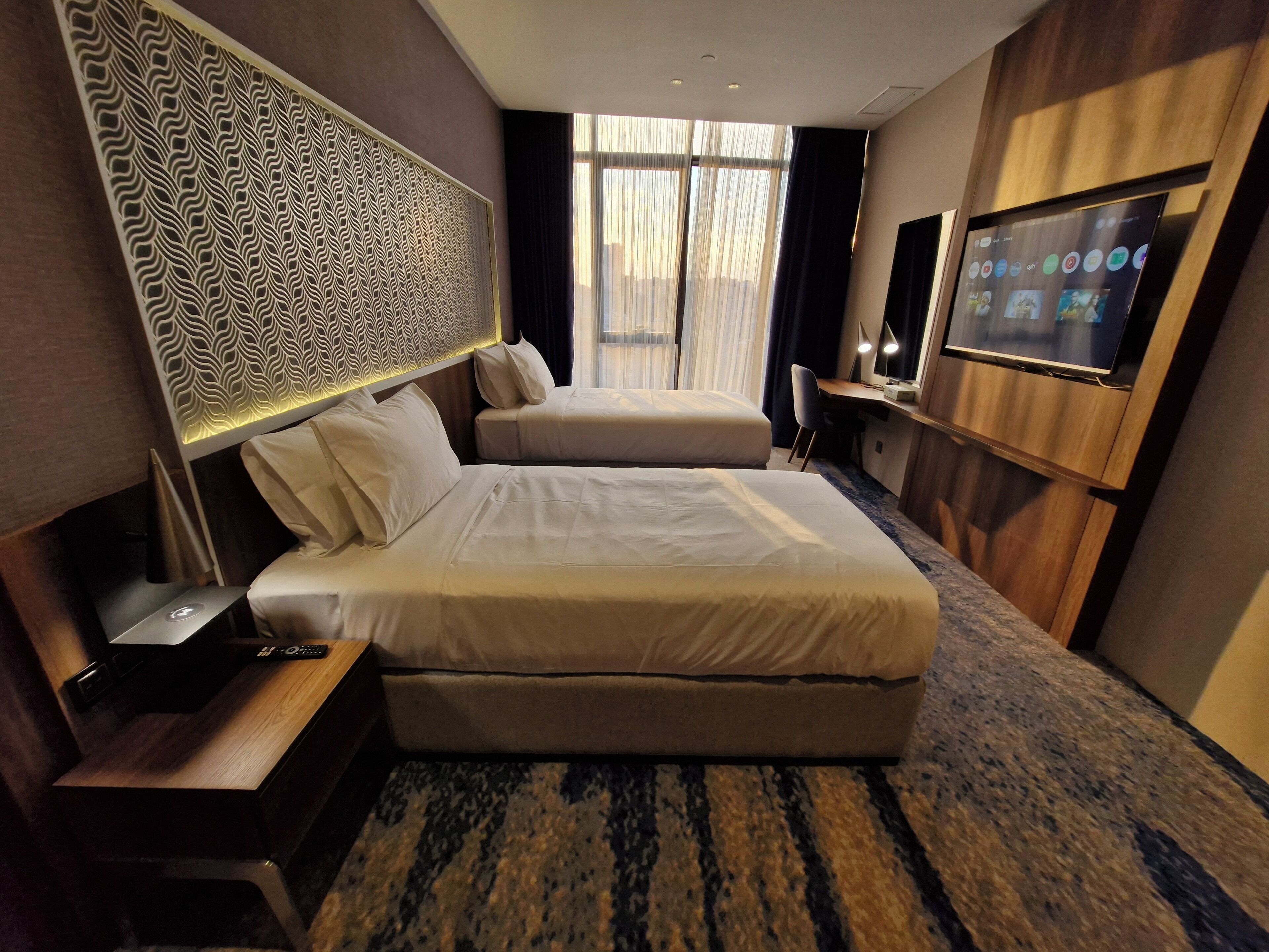 A hotel room with two beds, a desk, a TV, and a patterned headboard.