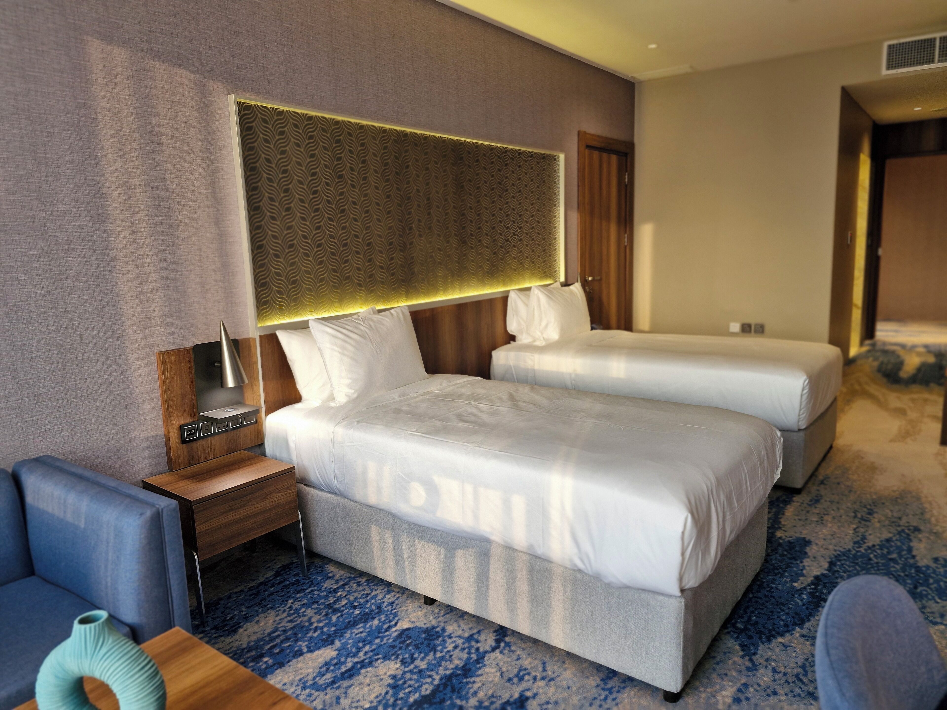 A hotel room with two beds, a nightstand, a lamp, a vase, and a blue patterned carpet.