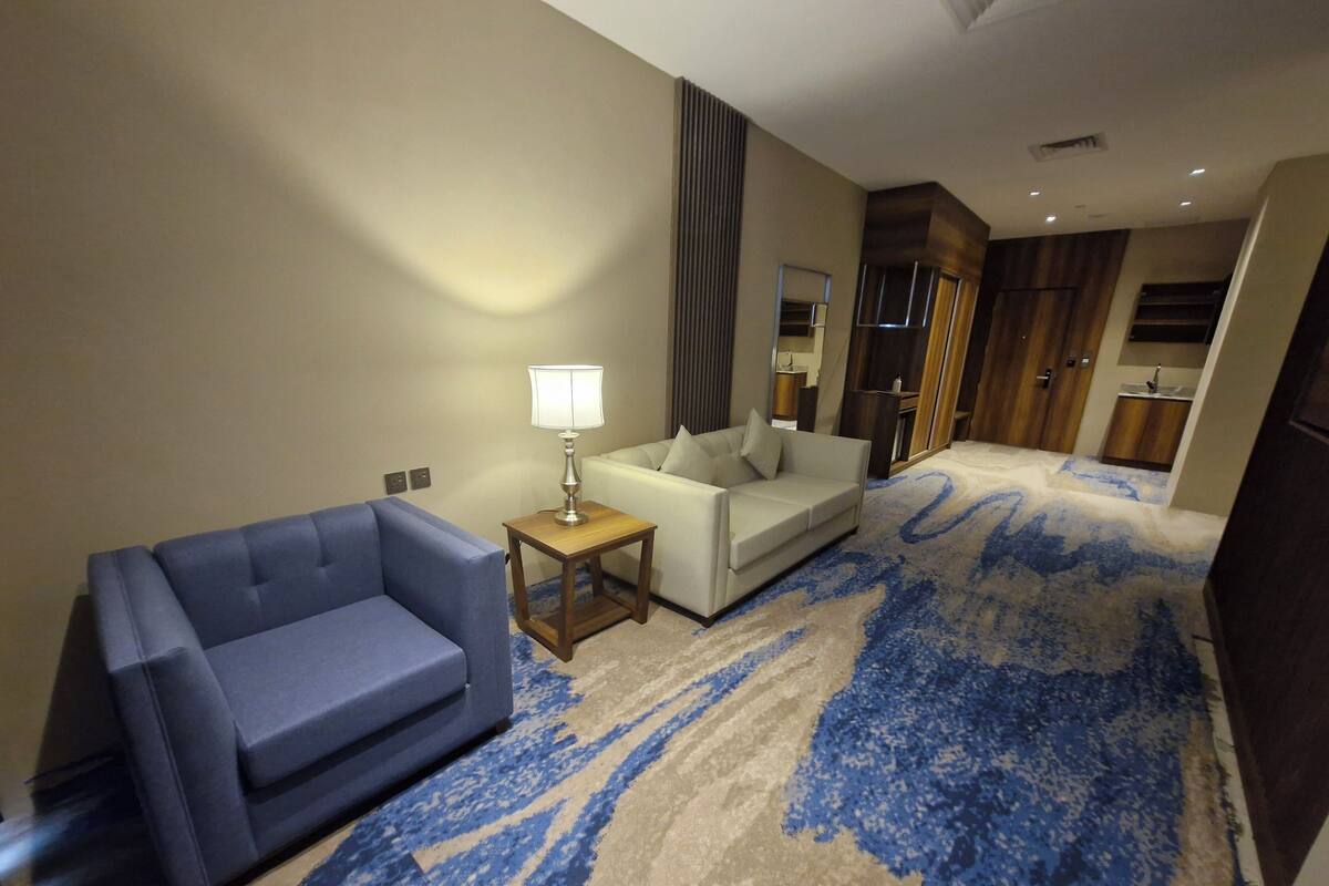 Une chambre d’hôtel avec un canapé bleu, un fauteuil blanc, une petite table en bois avec une lampe, une grande moquette à motifs et, au fond, une kitchenette.