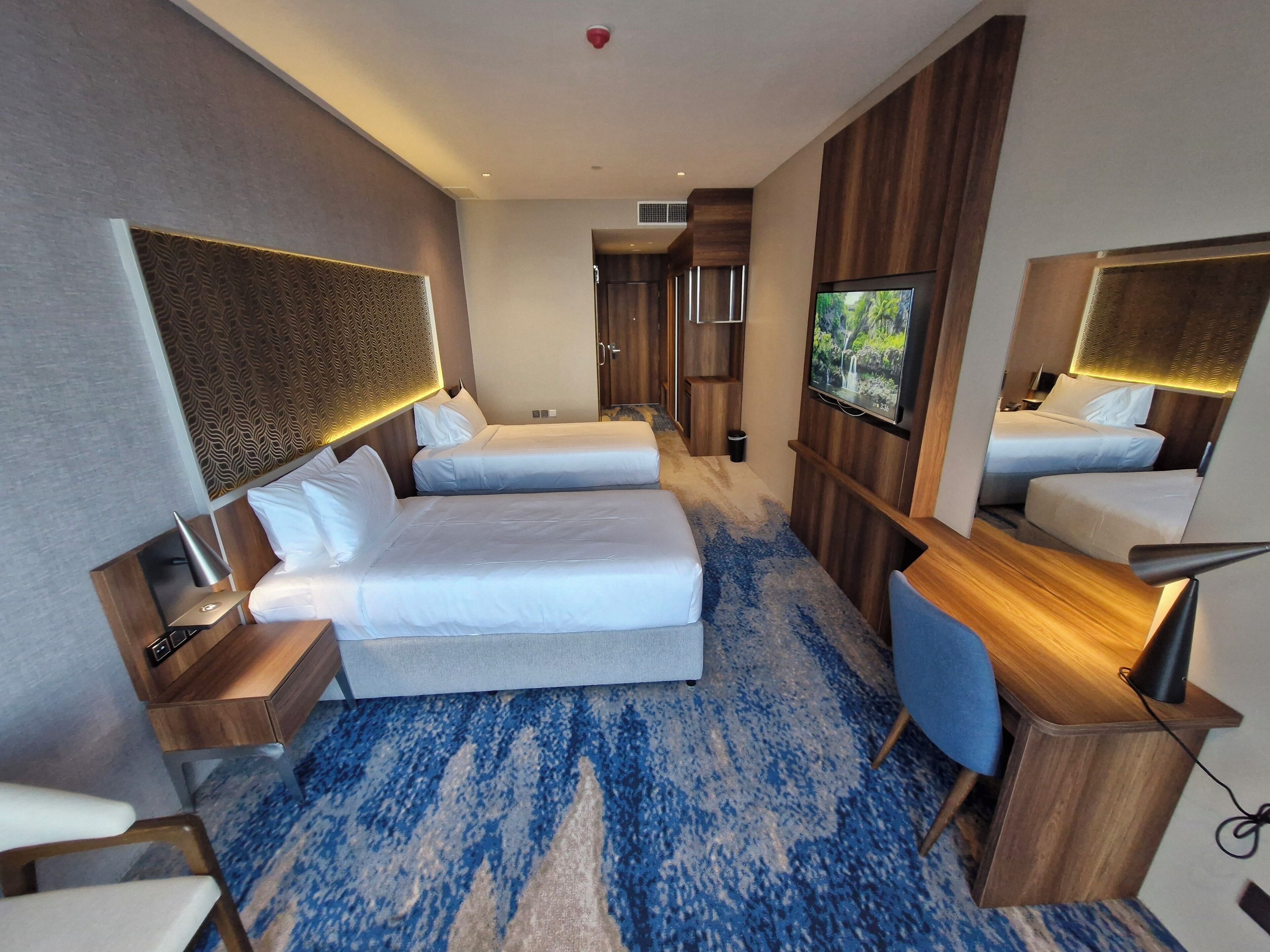 A hotel room with two beds, a desk, a chair, a TV, and a large mirror.