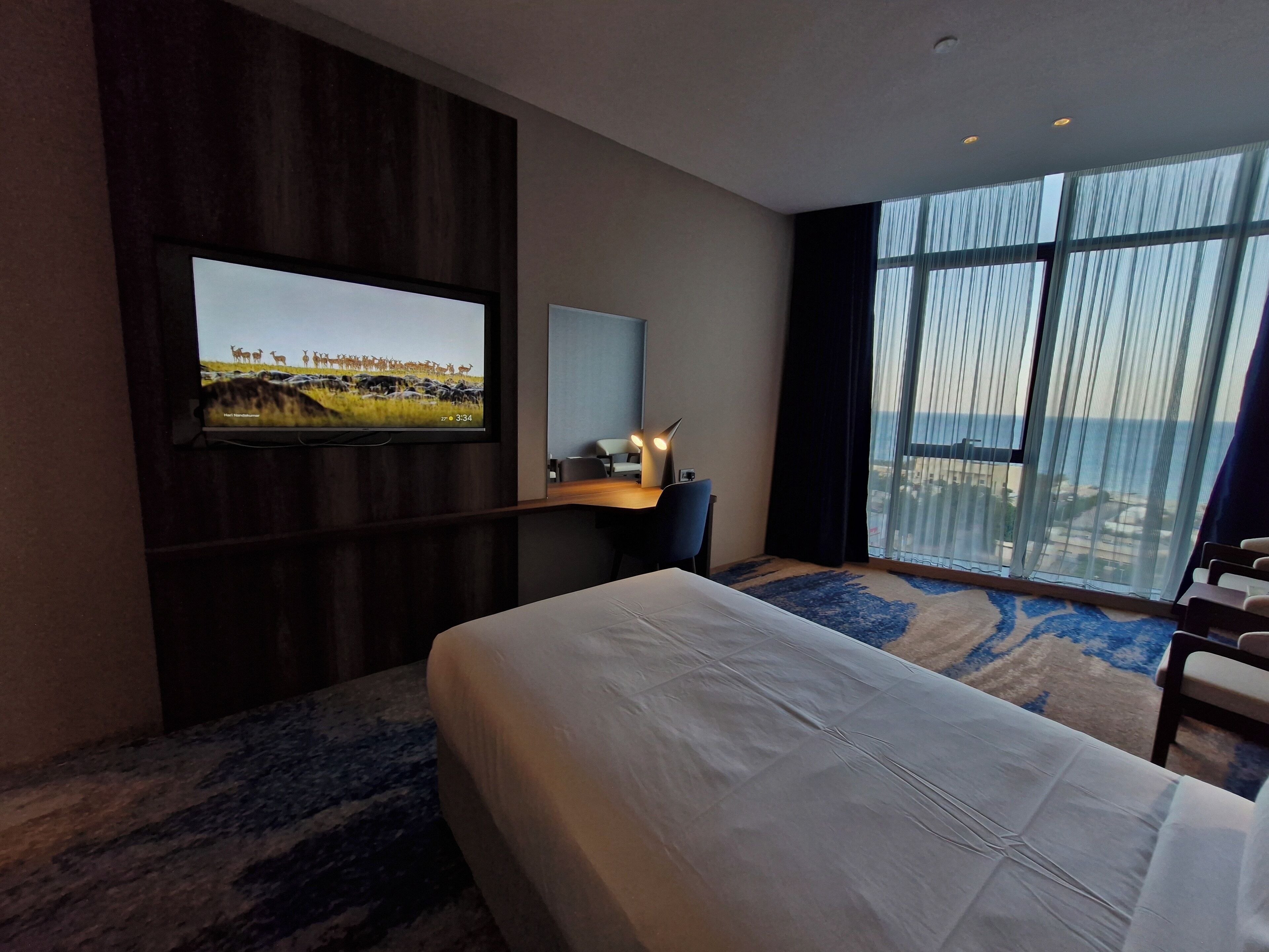 A hotel room with a large bed, a desk with a chair, a TV mounted on the wall, and a large window with a view of the beach.