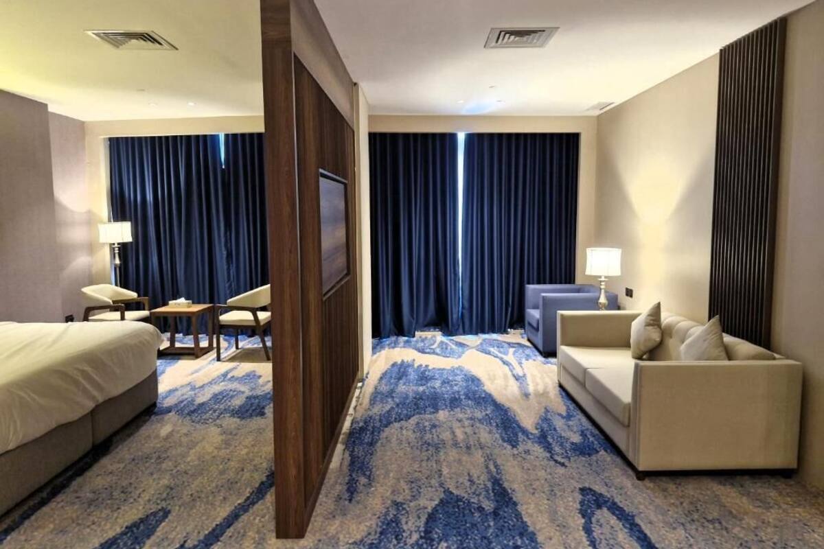 Une chambre d’hôtel avec un lit, un canapé, un bureau et une chaise. Il y a une grande fenêtre avec des rideaux, un téléviseur fixé au mur et une moquette à motifs.