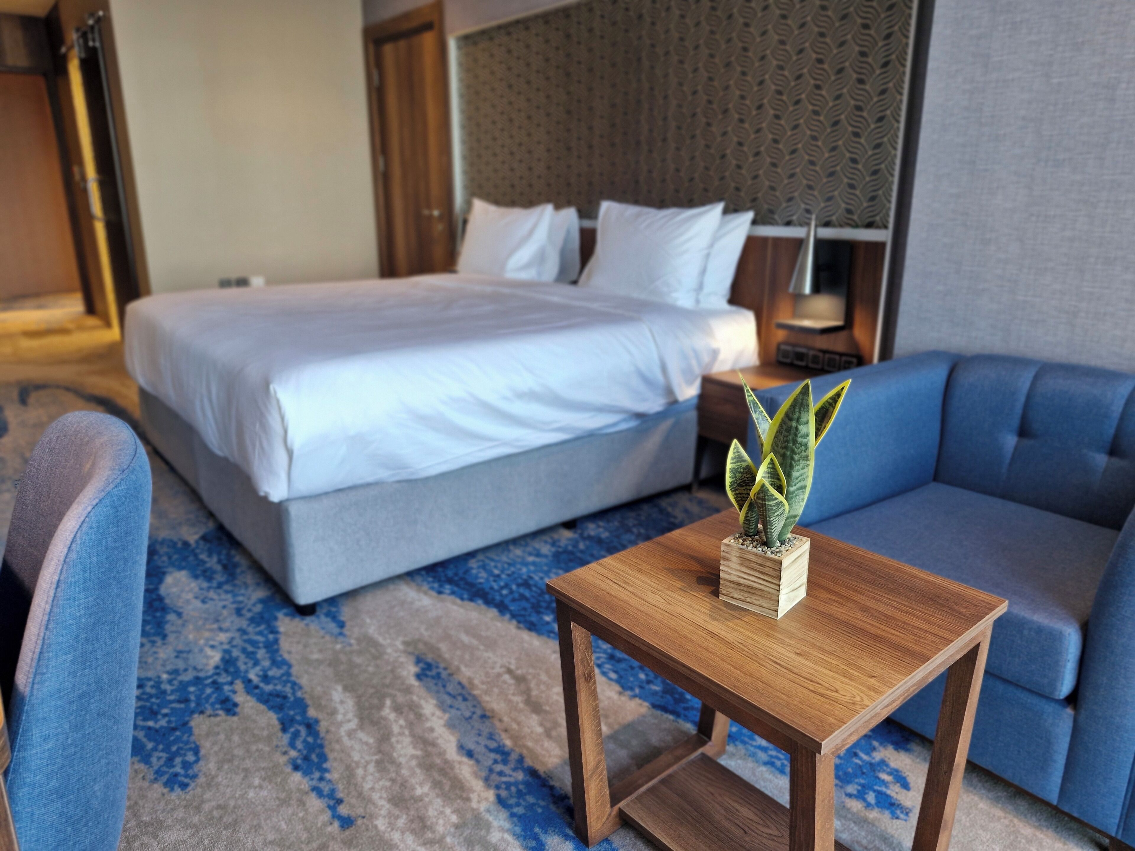 Ein Hotelzimmer mit einem Bett, einem blauen Sofa, einem Holztisch mit einer Pflanze und einem gemusterten Teppich.