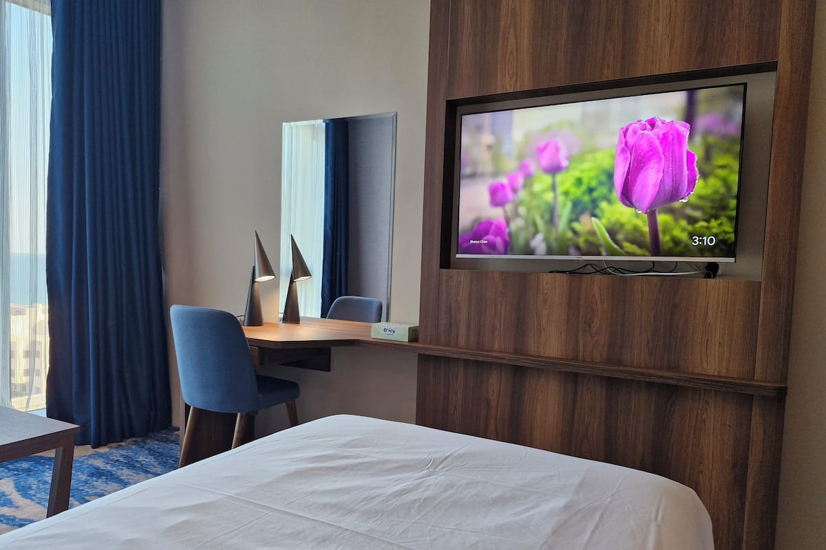 Une chambre d’hôtel comprenant un lit, un bureau avec une chaise, un téléviseur fixé au mur et une fenêtre avec des rideaux.