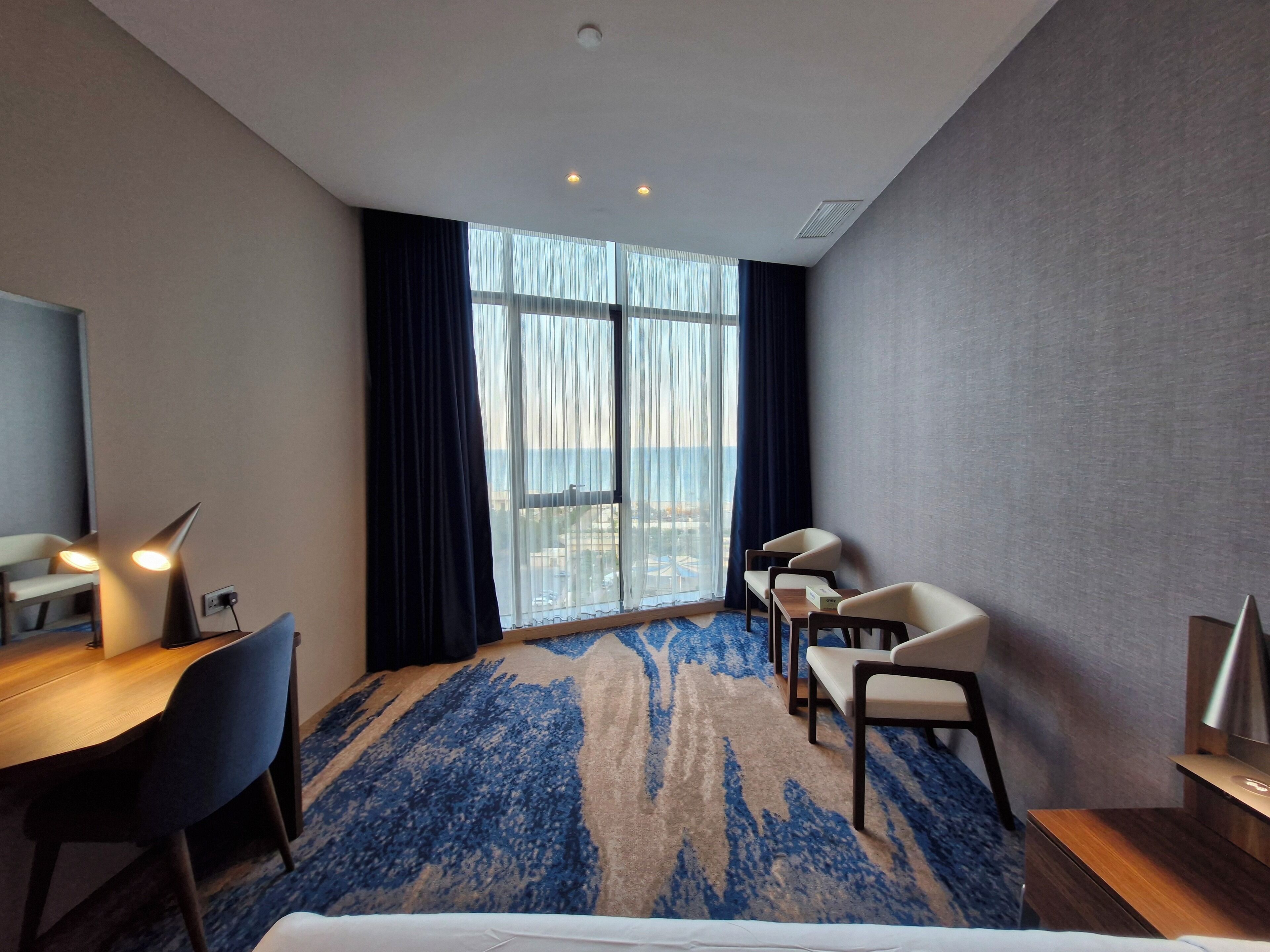 A hotel room with a large window, a desk with a chair, a bed, and a small table.
