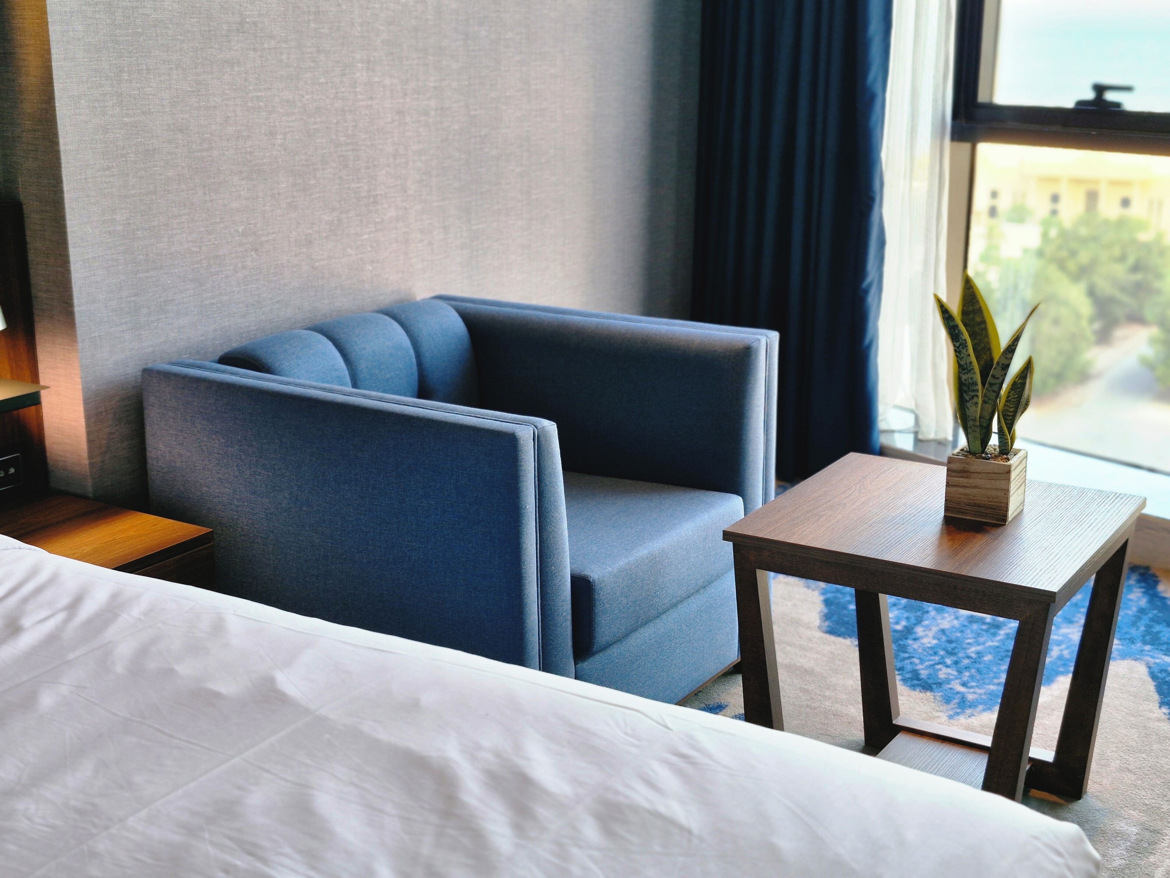 A hotel room with a bed, a blue armchair, a small wooden table with a potted plant, and a view of the outside through a window.
