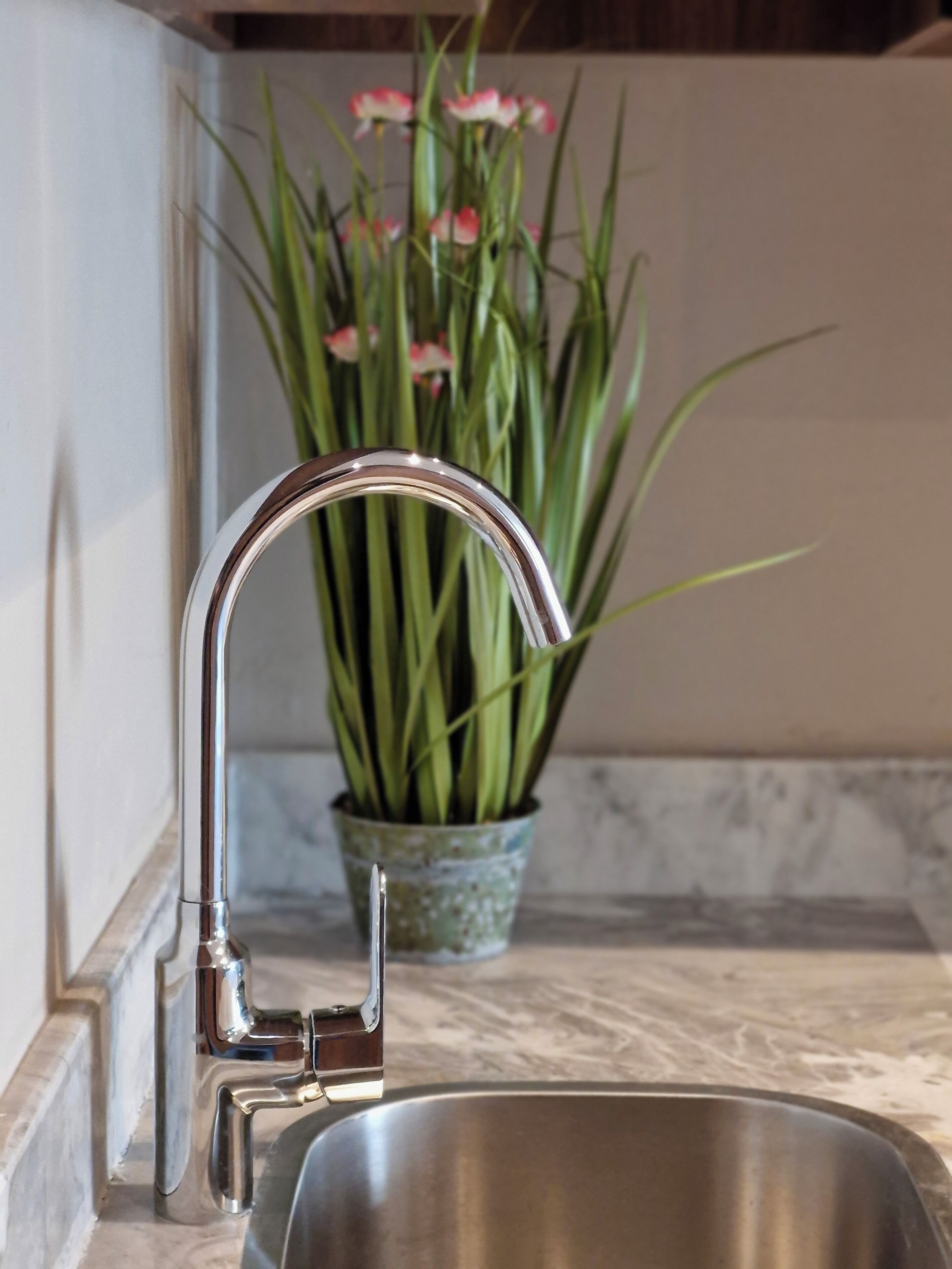 A stainless steel kitchen sink with a modern faucet, a marble countertop, and a potted plant with green leaves and pink flowers in the background.