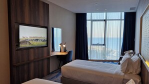 Et hotelværelse med to senge, et skrivebord med en computer, et fjernsyn på væggen og et stort vindue med havudsigt.