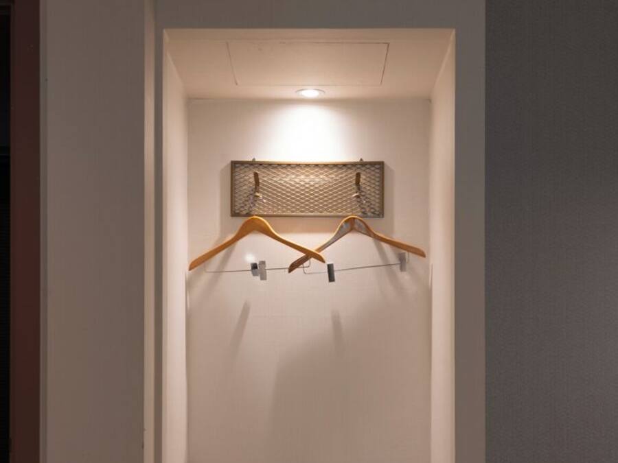 A compact hotel room with a small bed, a bedside table, a towel and a pillow on the bed, a hanging rack with clothes, and a wall-mounted shelf.