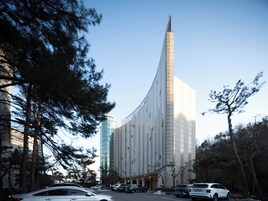 Un edificio moderno con un diseño curvo distintivo, rodeado de árboles y coches estacionados.