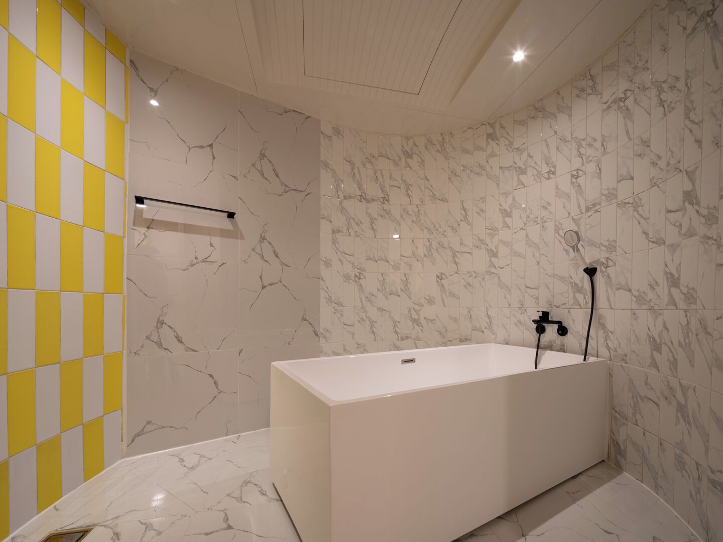 A modern bathroom with a marble floor and walls, a white bathtub, and a yellow and white checkered wall.