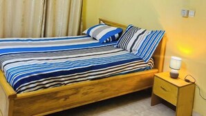 A bedroom with a wooden bed, striped bedding, a nightstand with a lamp, an air conditioner, and a painting on the wall.
