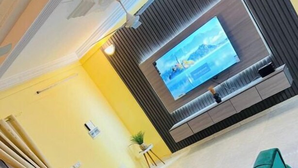 A living room with a large flat-screen TV mounted on a wall, green upholstered sofas, a small round coffee table, and a ceiling fan.