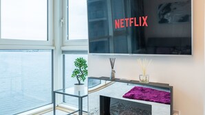 Een moderne woonkamer met een grote flatscreen televisie aan de muur, een glazen salontafel, een kleine plant en een open haard met een paars vloerkleed.