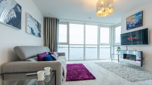 Een moderne woonkamer met een grijze bank, een bijzettafeltje, een open haard en een groot raam met uitzicht op het water.