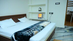 A hotel room with a bed, a bedside heater, a shelf with a kettle, and a bathroom area with a sink and mirror. - Shraddha Guest House (Ayodhya)