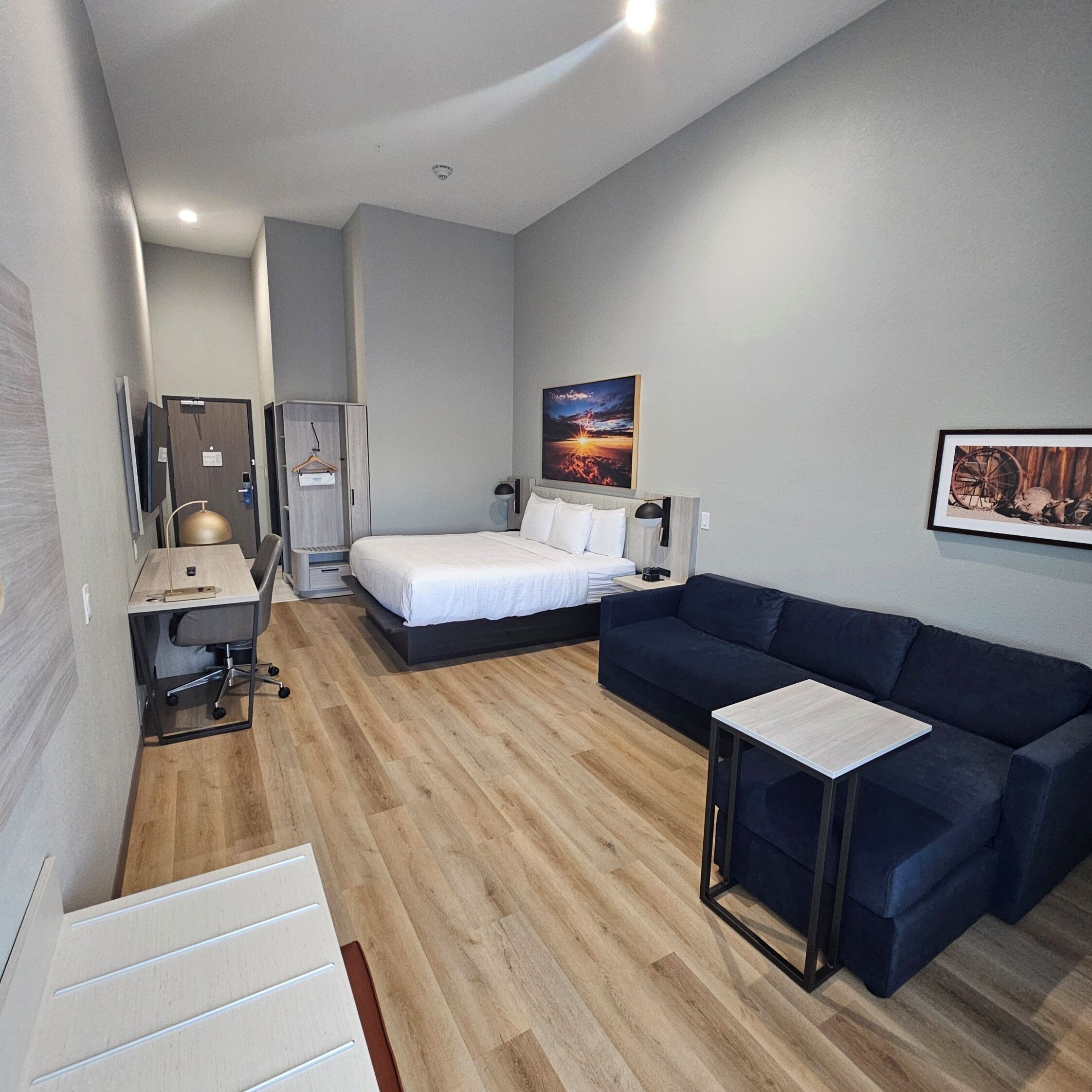 Una habitación de hotel moderna con una cama, un sofá azul oscuro, un escritorio con una silla, una mesita y un cuadro en la pared.