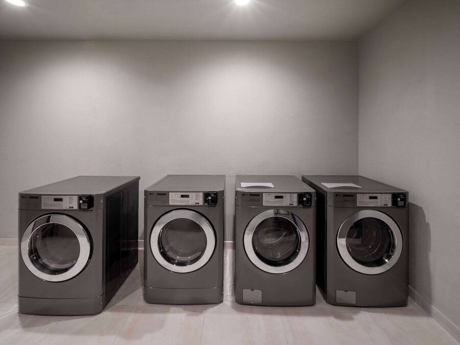 A row of four identical front-loading washing machines in a room with a plain white wall and a light-colored floor.